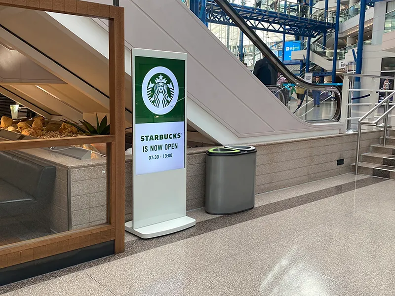 Freestanding digital poster beside an escalator in a shopping centre